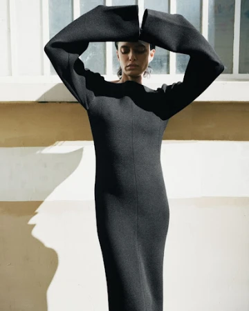 Model in a long black dress with arms raised overhead, set against an exterior wall with striped shadows.