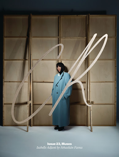 A magazine cover features a full-length portrait of a woman in a blue coat, with a light wooden frame and large handwritten text overlay.