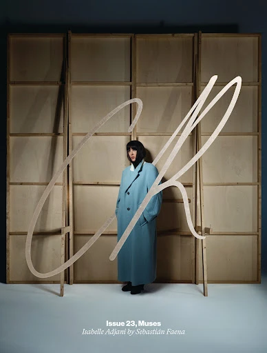 A magazine cover features a full-length portrait of a woman in a blue coat, with a light wooden frame and large handwritten text overlay.
