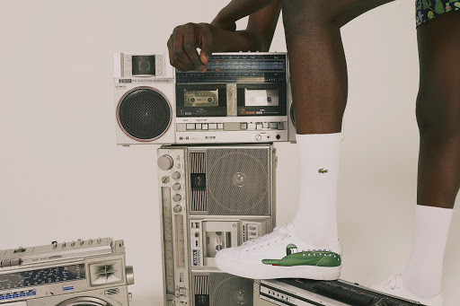 Person stands with retro boomboxes, white Lacoste sneakers, and white socks with Lacoste logo; studio lighting; neutral background.