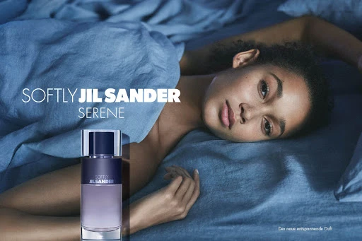 Adult female model poses on blue bedding next to a Jil Sander perfume bottle in a beauty advertisement with soft lighting.
