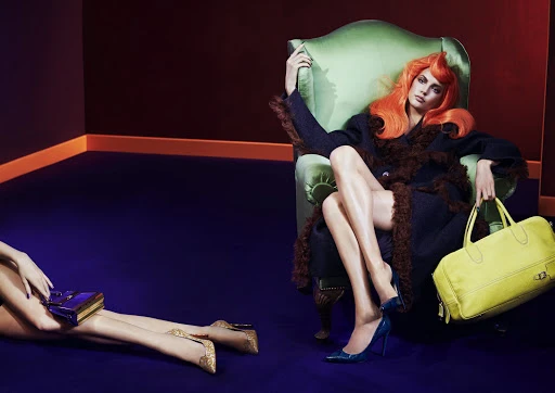 Fashion shot of a red-haired woman in a green chair holding a yellow bag, with another woman reclining on a blue carpet nearby.