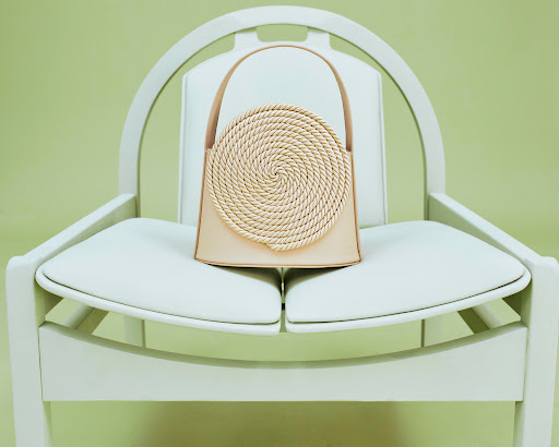 Beige leather handbag with woven straw circle detail, sitting on a modern white chair against a soft pale green background.