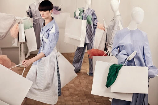 Woman in blue and white striped dress surrounded by shopping bags and mannequins in a fashion store setting with natural light.