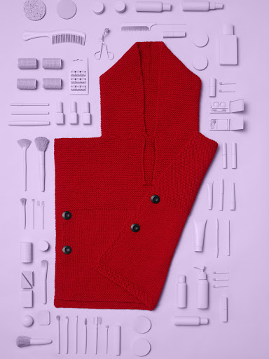 Flat lay of a red knit hooded poncho surrounded by assorted white beauty and makeup products on a light purple backdrop.