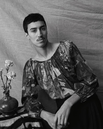 Monochrome portrait of a man with a mustache wearing a patterned long-sleeve blouse next to a vase of flowers.