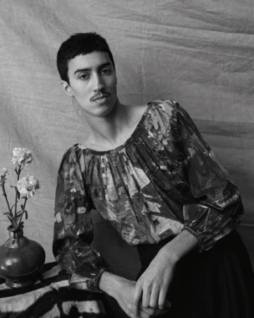 Monochrome portrait of a man with a mustache wearing a patterned long-sleeve blouse next to a vase of flowers.