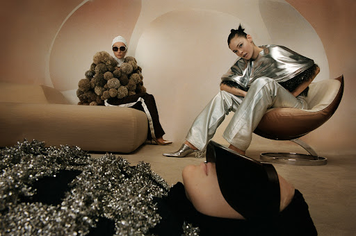Two women pose in a studio with futuristic fashion. One sits on a cream chair in a silver outfit, the other on a couch covered in ball shapes.