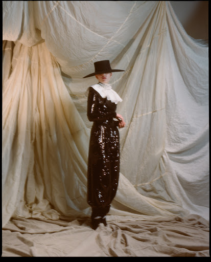 Woman in a sparkly black Pilgrim-style dress with a ruffled collar and a wide-brimmed hat, posing in front of draped fabric.
