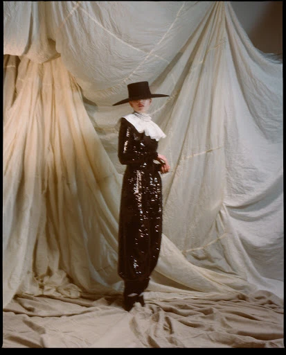 Woman in a sparkly black Pilgrim-style dress with a ruffled collar and a wide-brimmed hat, posing in front of draped fabric.
