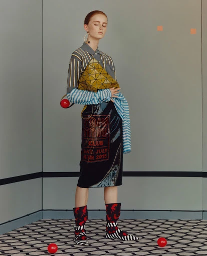 Fashion shot of a red-haired woman in a striped shirt, patterned skirt, and matching boots, holding a geometric bag in an indoor setting.