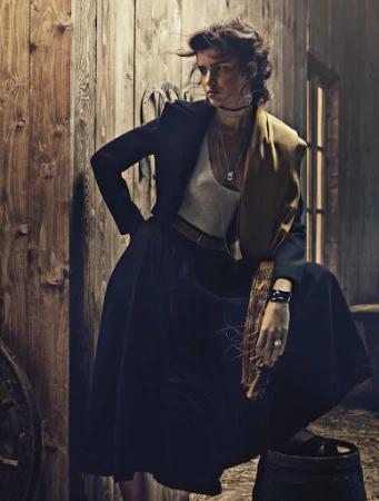 Model in dark jacket, white top, and long skirt leaning against a wooden barn wall styled by Sylvain Cabouat