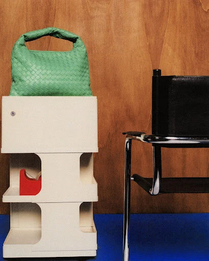 A woven green handbag is placed on a white tiered display unit, next to a black chair with a chrome frame against a wood and blue backdrop.