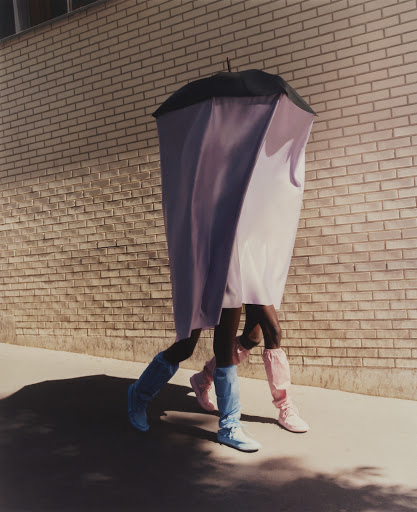 Two people walking side-by-side, one wearing a pale purple covering and the other wearing bright blue boots against a brick wall.