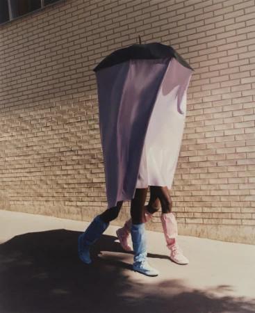 Two people walking side-by-side, one wearing a pale purple covering and the other wearing bright blue boots against a brick wall.