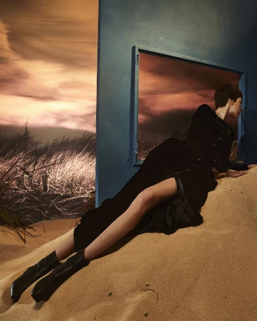 Model wearing black dress reclining on a sand dune next to a blue wall with a framed window showing a cloudy sunset landscape.