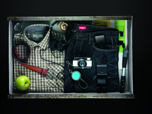 Overhead studio shot of a clothing and accessory collection in a metal drawer featuring a plaid shirt, jeans, and various gadgets.