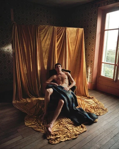 Shirtless male model poses in an armchair draped with blue fabric, set against a gold curtain backdrop and wallpapered room with natural light.