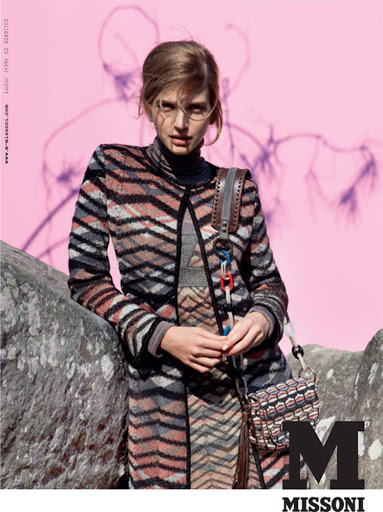 A woman poses in a patterned knit coat and dress, accessorized with a shoulder bag, against a vibrant pink backdrop.