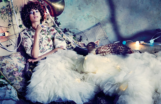Woman in floral blouse and white ruffled skirt poses with a fawn on a set with pillows, string lights, and a sousaphone.
