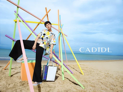 A woman in a patterned top and black pants poses on a beach with colorful wooden sticks, holding a tote bag in a fashion-forward shot.
