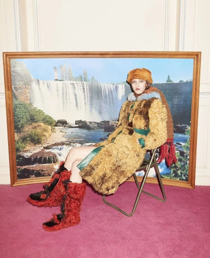 A woman sits on a metal folding chair in front of a painting of a waterfall wearing a fuzzy hat, coat, and boots indoors.
