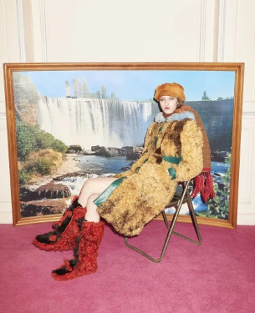 A woman sits on a metal folding chair in front of a painting of a waterfall wearing a fuzzy hat, coat, and boots indoors.