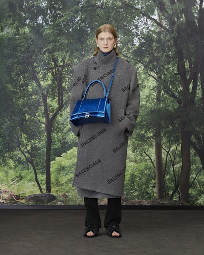 Fashion model poses in a patterned grey coat with a blue handbag against a green forest backdrop for a Balenciaga campaign.