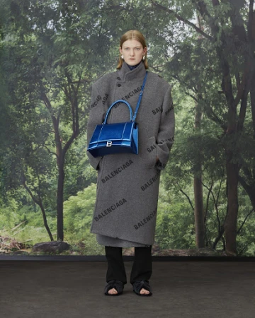 Fashion model poses in a patterned grey coat with a blue handbag against a green forest backdrop for a Balenciaga campaign.