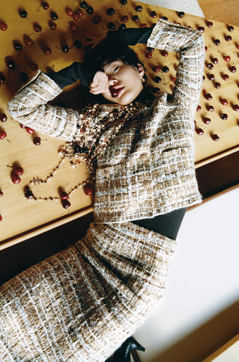 A woman in a tweed skirt and jacket lies on a wooden surface surrounded by cherries, looking at the camera with a cherry in her mouth.