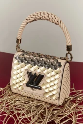 Beige handbag with gold and silver scale appliqués and a braided top handle resting on a pile of gold cords