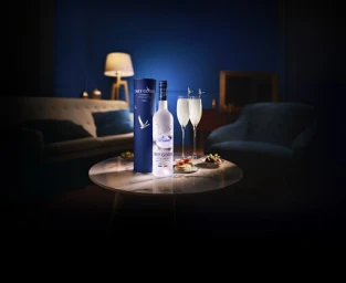Grey Goose vodka bottle and canister with two champagne glasses on a marble coffee table in a dimly lit living room setting.