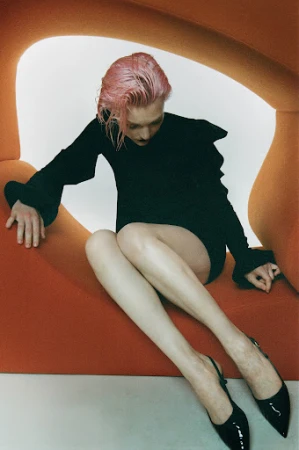 Woman with pink wet-look hair wearing a black dress posing within a curved orange sculptural furniture piece.