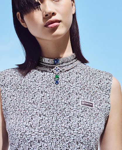 Asian woman poses in a sleeveless grey tweed dress with a luxury diamond and gemstone necklace against a bright blue background.