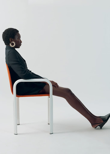 Elegant Black woman with short natural hair wearing blazer dress and slingbacks, sitting sideways on orange chair against white backdrop.
