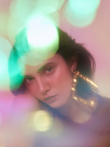 Close-up of a woman with fair skin, adorned with large, gold earrings, posing in a colorful, bokeh-filled studio environment.