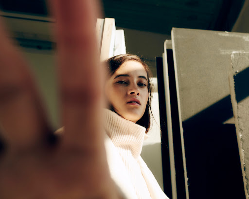 Portrait of a young woman in a cream turtleneck sweater with hand in foreground, set against a neutral studio backdrop.