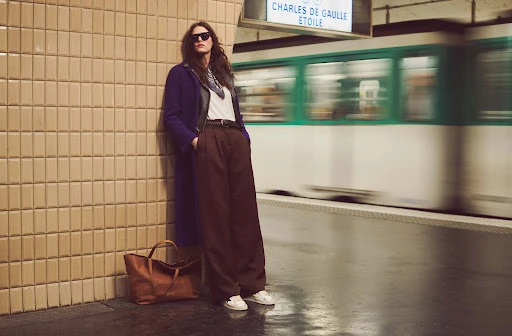 A woman in a long purple coat, brown pants, and sunglasses leans against tiled wall in a subway station with a blurred train.