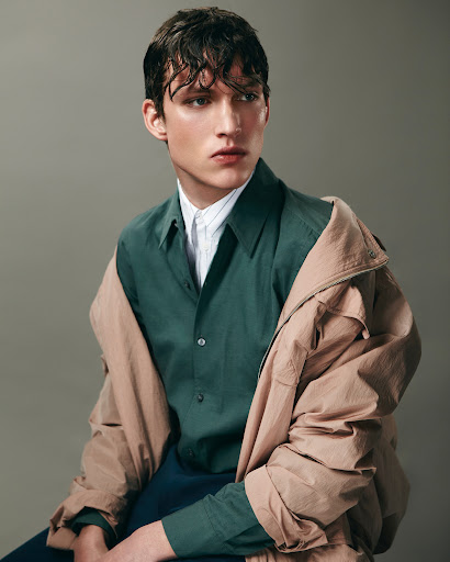 Studio shot of a young adult man wearing a green button-down shirt and jacket, looking off camera, with a gray background.