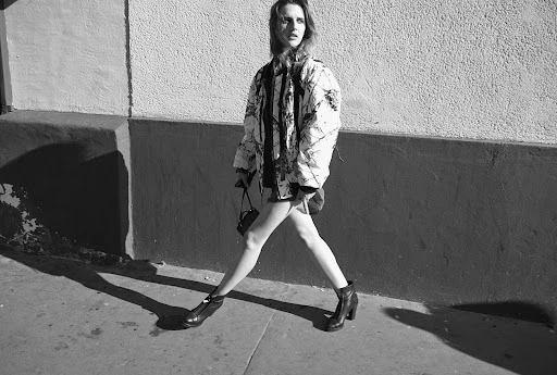 A woman in a patterned quilted jacket walks past a textured wall with her shadow cast behind her.