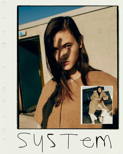 Portrait of a young woman with long dark hair, blocking the sun with her hand, wearing a beige top in front of a concrete wall.