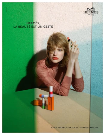 Fashion shot: Woman poses with Hermès lipstick in front of a color-blocked background with shades of green, beige and white, studio shot.