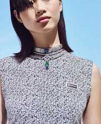 Close-up portrait of a woman wearing layered diamond necklaces and a patterned top against a clear blue sky background.