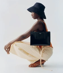 Model in a black bucket hat wearing an orange top and holding a structured black handbag against a white background
