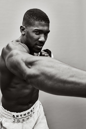 Monochrome portrait of shirtless male boxer Anthony Joshua in a fighting stance, wearing white shorts with his name on the waistband.