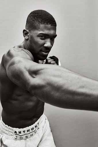 Monochrome portrait of shirtless male boxer Anthony Joshua in a fighting stance, wearing white shorts with his name on the waistband.