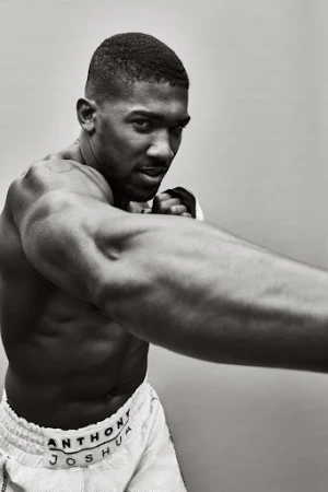 Monochrome portrait of shirtless male boxer Anthony Joshua in a fighting stance, wearing white shorts with his name on the waistband.