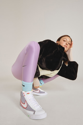 Model in a crouching pose wearing purple leggings, a black furry jacket, and high-top sneakers on a white background