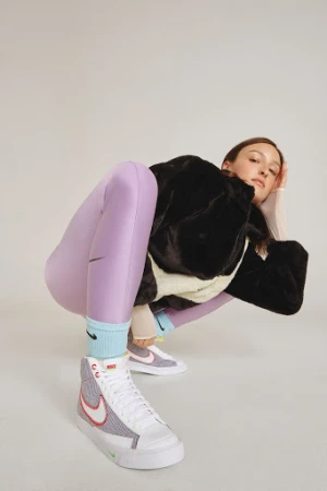 Model in a crouching pose wearing purple leggings, a black furry jacket, and high-top sneakers on a white background