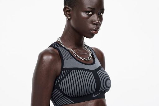 Close-up shot of a Black woman with short hair, wearing a gray and black sports bra and silver necklaces, posing against a white background.